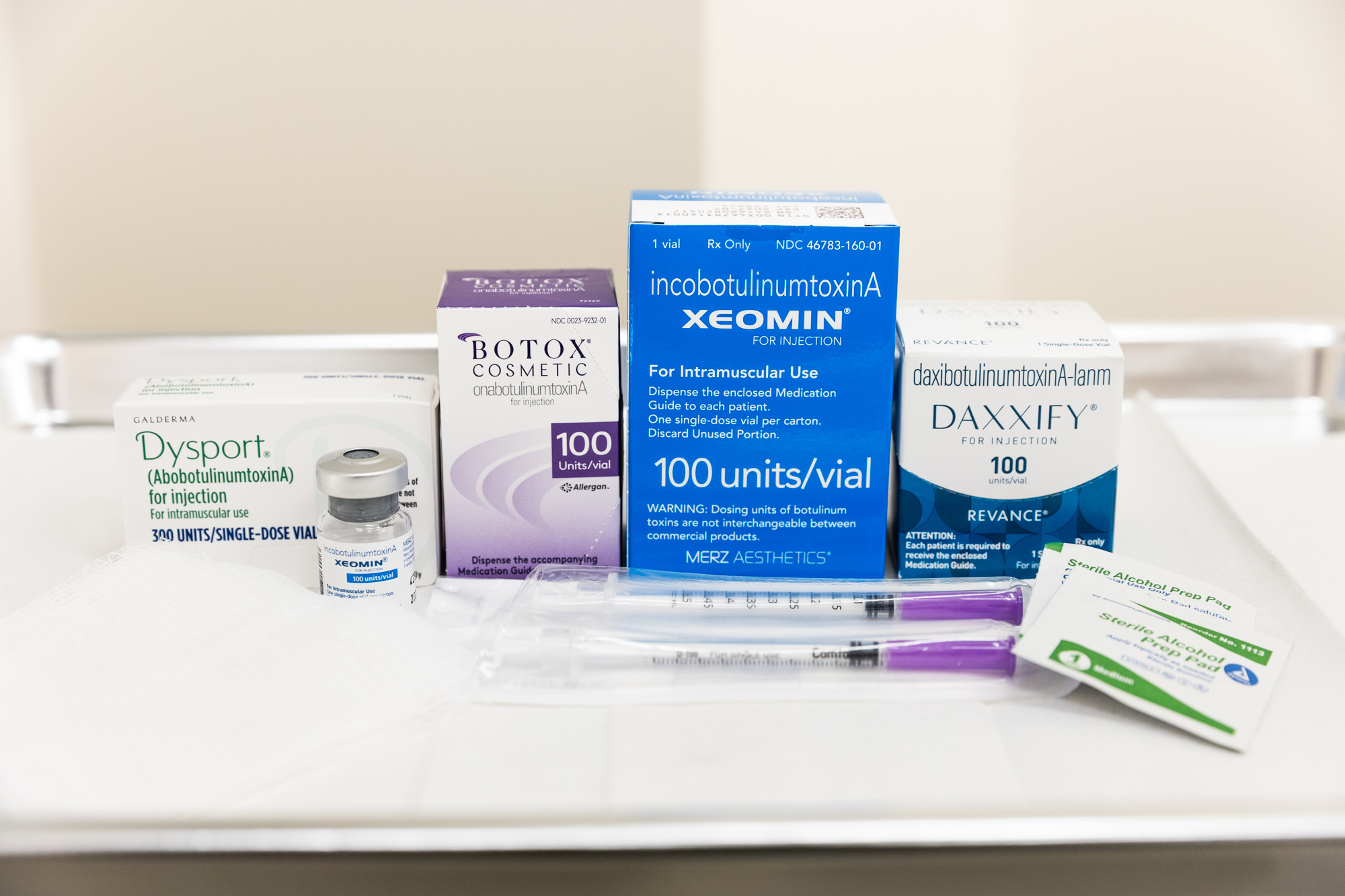 Several different brands of wrinkle relaxers, including Xeomin, Botox, and Dysport, sit on a white counter, along with injection syringes and cleansing pads. When choosing between Botox and fillers in Brooklyn Park, it's important to consider the variety of products available before making your decision.