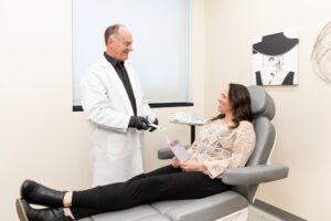 Enrichment Skin Solutions co-founder Michael Johnson, wearing a white lab coat, black collared shirt, and black gloves, discusses ezGEL near Maple Grove with a female patient lying on a treatment bed. This treatment utilizes platelets, plasma, and fibrin in the blood to rejuvenate skin.