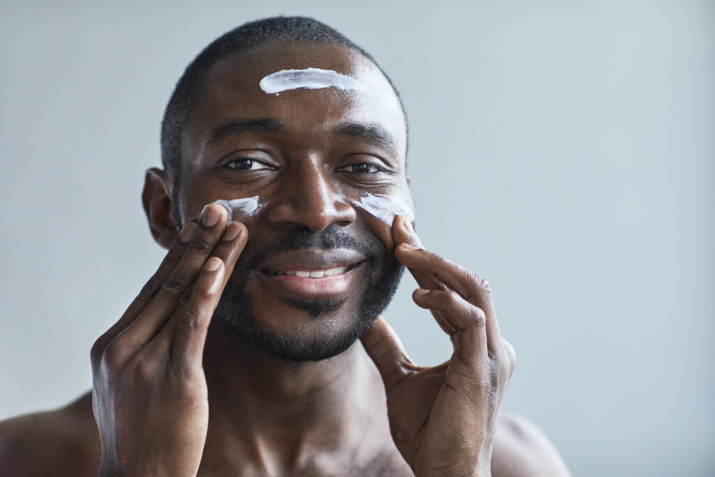 A young black man applying a moisturizing cream o his skin after visiting the team at Enrichment Skin Solutions for microneedling near Maple Grove. The cream will help his skin become smoother and healthier as it heals.